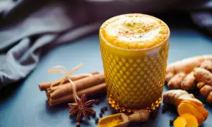 Ein goldgelbes Getränk mit Kurkuma in Kaffee, verfeinert mit Zimt und Sternanis. Daneben liegen frische Kurkumawurzeln und eine kleine Holzschaufel mit Kurkumapulver.