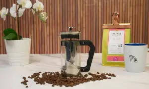 French press mit Kaffeesatz, bereit zum Entsorgen. Daneben Kaffeebohnen, eine Kaffeetüte und eine Tasse.