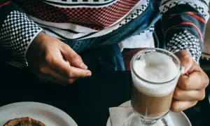 Person hält ein Glas mit schaumigem, kaltem Cappuccino. Auf dem Tisch liegt ein kleines Gebäck.