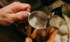 Hände halten Kännchen, während Dampf für perfekten Milchschaum wie aus der French Press aufsteigt.