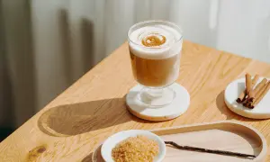 Ein cremiger Zimt Latte mit aufgeschäumter Milch und Zimtdekor. Daneben brauner Zucker, Zimtstangen und eine Vanilleschote.