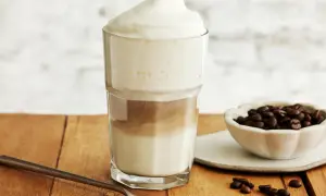 Ein hoher Latte Macchiato Eiskaffee mit Milchschaum, daneben eine Schale Kaffeebohnen und ein Löffel.
