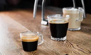 Zwei Gläser mit Kaffee stehen auf einem Holztisch. Ein kleiner starker Kaffee mit Crema im Vordergrund, daneben ein weiterer Kaffee und im Hintergrund ein Milchkaffee.