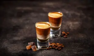 Zwei geschichtete Kaffeegetränke in Gläsern, umgeben von Kaffeebohnen und braunem Zucker. So kann man mit Kaffeebohnen köstliche Drinks zaubern.