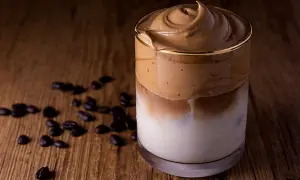 Ein cremiger Dalgona Kaffee ohne Zucker, serviert in einem Glas auf einem Holztisch, umgeben von Kaffeebohnen.