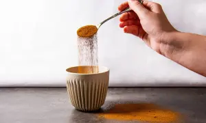 Eine Hand schüttet mit einem Löffel braunes Pulver in eine gerippte Tasse. Der Pulverregen erinnert an instant kaffee koffein.