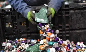 Hände in Handschuhen kippen bunte Nespresso-Kapseln aus einer grünen Tüte in einen schwarzen Behälter. Die fachgerechte Entsorgung von Nespresso-Kapseln ist wichtig.
