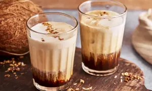 Zwei Gläser mit cremigem Kaffee mit Kokosmilch, garniert mit Kokosraspeln. Daneben eine Kokosnuss.