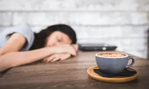 Eine Frau schläft auf dem Tisch, ihr Kopf ruht auf ihren Armen. Neben ihr steht eine Tasse Kaffee, die sie wohl abends trinken wollte.