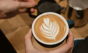 Perfekte Cappuccino Zubereitung: Eine Hand hält die Tasse mit Latte Art, während im Hintergrund Milch aufgeschäumt wird.
