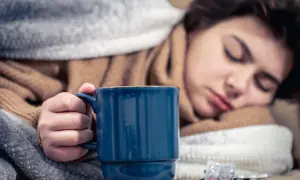 Müde Frau mit Fieber hält eine Tasse Kaffee, um sich aufzuwärmen. Medikamente liegen daneben.