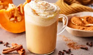 Ein cremiger Pumpkin Spice Latte mit Sahne und Zimt, daneben Kürbisspalten, Zimtstangen und Gewürznelken.
