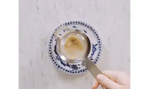 Hand hält Sieb mit braunem Schaum über blau-weißem Teller. Ein leckerer soy latte wird zubereitet.