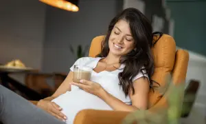 Glückliche Schwangere genießt einen Kaffee. Sie sitzt entspannt in einem Sessel und hält ein Glas mit einem cremigen Getränk.