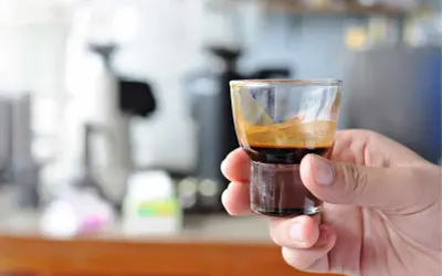 Eine Hand hält ein kleines Glas mit einem dunklen, cremigen Ristretto. Die Zubereitung ist perfekt, die Crema glänzt.