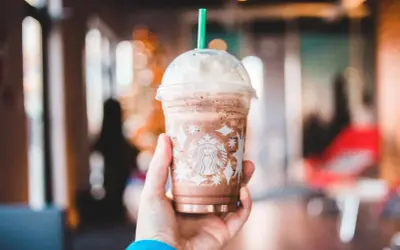 Eine Hand hält einen Schokoladen-Frappuccino von Starbucks, der mit Schlagsahne und einem grünen Strohhalm gekrönt ist. Perfekt für heiße Tage, diese Starbucks Rezepte Frappuccino sind ein Genuss.