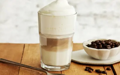 Ein hoher Latte Macchiato Eiskaffee mit Milchschaum, daneben eine Schale Kaffeebohnen und ein Löffel.