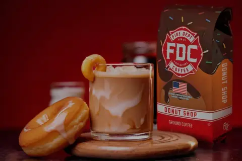 Ein cremiger iced americano mit einem Donut-Garnitur, daneben ein Donut und eine Packung Fire Dept. Coffee.