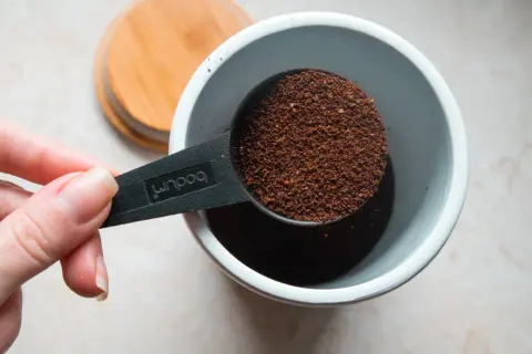 Eine Hand hält einen Löffel mit frisch gemahlenem Kaffeepulver, bereit, es in einen Behälter zu geben. Die Frage ist: wie viel Kaffeepulver für den perfekten Morgen?
