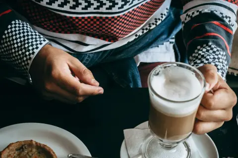 Person hält ein Glas mit schaumigem, kaltem Cappuccino. Auf dem Tisch liegt ein kleines Gebäck.