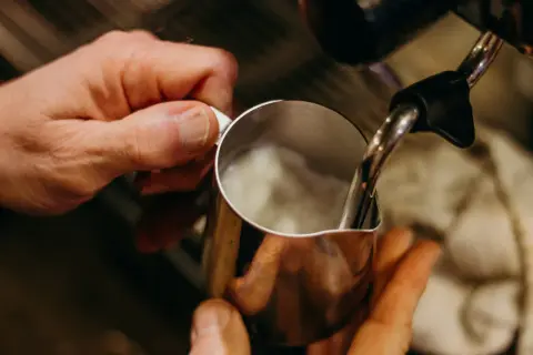 Hände halten Kännchen, während Dampf für perfekten Milchschaum wie aus der French Press aufsteigt.