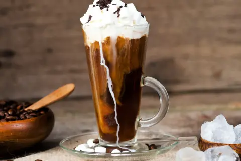 Ein erfrischender iced espresso mit Sahne, Marshmallows und Schokostreuseln, daneben Kaffeebohnen und Eiswürfel.