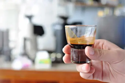 Eine Hand hält ein kleines Glas mit einem dunklen, cremigen Ristretto. Die Zubereitung ist perfekt, die Crema glänzt.
