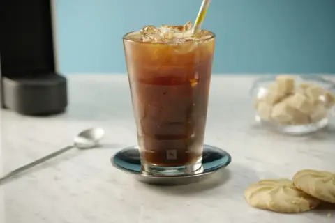 Erfrischender Nespresso Iced Coffee mit Eiswürfeln und einem bunten Strohhalm, daneben Kekse und Zuckerwürfel.