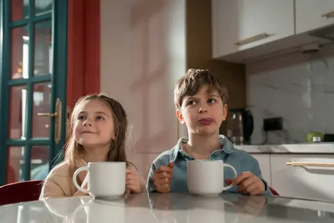 Zwei Kinder mit Tassen, die sich fragen, ab welchem Alter man Kaffee trinken darf. Der Junge leckt sich die Lippen.