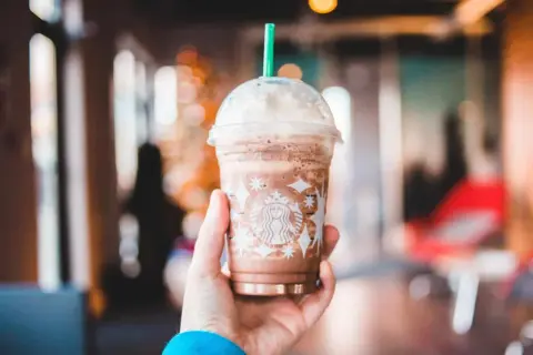 Eine Hand hält einen Schokoladen-Frappuccino von Starbucks, der mit Schlagsahne und einem grünen Strohhalm gekrönt ist. Perfekt für heiße Tage, diese Starbucks Rezepte Frappuccino sind ein Genuss.