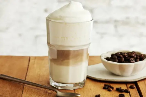 Ein hoher Latte Macchiato Eiskaffee mit Milchschaum, daneben eine Schale Kaffeebohnen und ein Löffel.