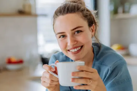 Lächelnde Frau hält eine Tasse Kaffee. Sie genießt den Moment, vielleicht trinkt sie alten Kaffee, um den Tag zu beginnen.