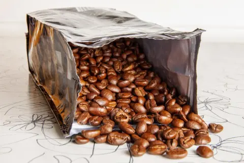 Ein offener Beutel mit gerösteten Kaffeebohnen, die auf eine gemusterte Oberfläche fallen. Die Frage, wie lange sind kaffeebohnen haltbar, beschäftigt jeden Kaffeeliebhaber.