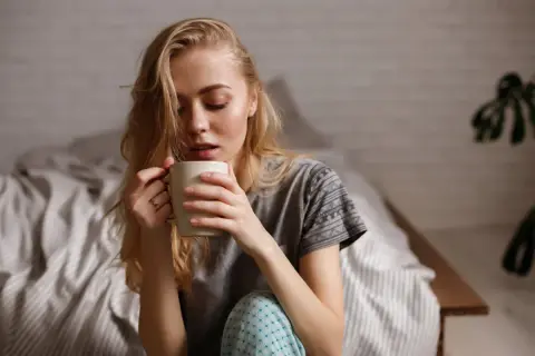 Junge Frau sitzt im Bett, hält eine Tasse und denkt nach. Ihr Kaffee schmeckt nicht mehr.