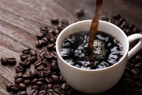 Heißer schwarzer Kaffee wird in eine weiße Tasse gegossen, umgeben von gerösteten Kaffeebohnen. Ein gesunder Start in den Tag.