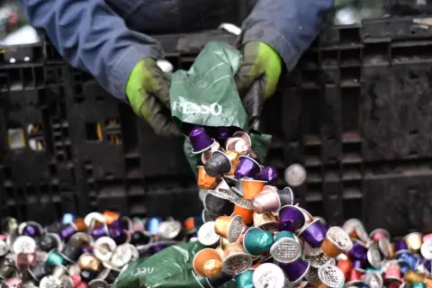 Hände in Handschuhen kippen bunte Nespresso-Kapseln aus einer grünen Tüte in einen schwarzen Behälter. Die fachgerechte Entsorgung von Nespresso-Kapseln ist wichtig.