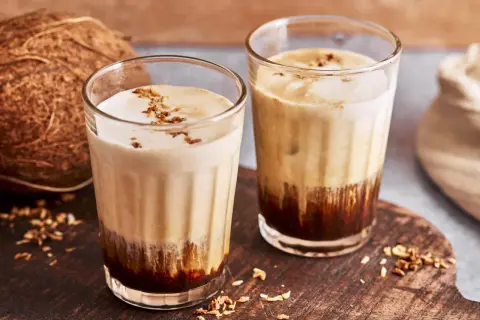 Zwei Gläser mit cremigem Kaffee mit Kokosmilch, garniert mit Kokosraspeln. Daneben eine Kokosnuss.
