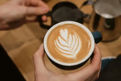 Perfekte Cappuccino Zubereitung: Eine Hand hält die Tasse mit Latte Art, während im Hintergrund Milch aufgeschäumt wird.