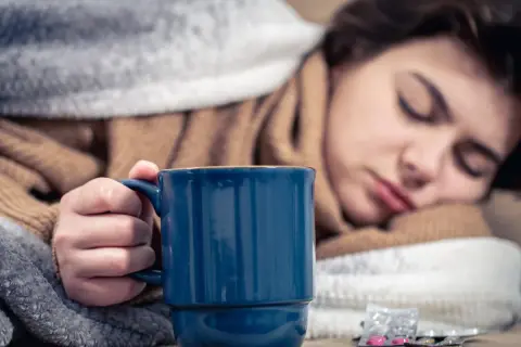 Müde Frau mit Fieber hält eine Tasse Kaffee, um sich aufzuwärmen. Medikamente liegen daneben.