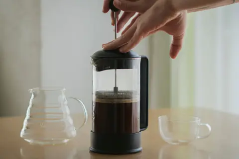 Hände drücken den Stempel einer French Press herunter, um Kaffee zu kochen. Daneben stehen eine leere Glaskaraffe und eine Tasse.