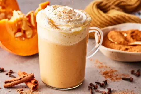 Ein cremiger Pumpkin Spice Latte mit Sahne und Zimt, daneben Kürbisspalten, Zimtstangen und Gewürznelken.