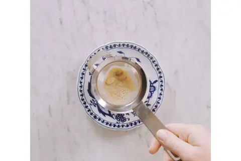 Hand hält Sieb mit braunem Schaum über blau-weißem Teller. Ein leckerer soy latte wird zubereitet.