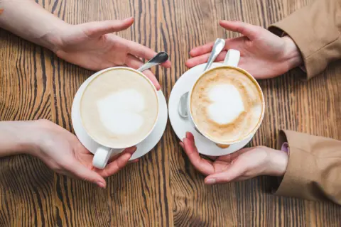 Zwei Hände reichen sich zwei Kaffees, ein Flat White und ein Cappuccino, mit Herz-Latte-Art auf Holztisch.