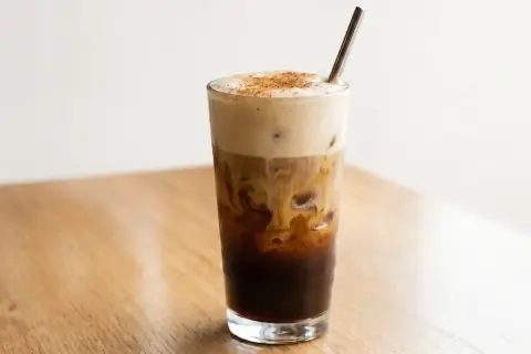 Ein erfrischender, geschüttelter Espresso mit cremiger Haube und Zimt bestreut, serviert in einem Glas mit Eiswürfeln und einem Metallstrohhalm.