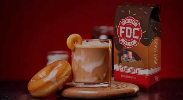 Ein cremiger iced americano mit einem Donut-Garnitur, daneben ein Donut und eine Packung Fire Dept. Coffee.