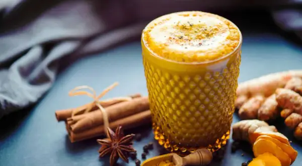 Ein goldgelbes Getränk mit Kurkuma in Kaffee, verfeinert mit Zimt und Sternanis. Daneben liegen frische Kurkumawurzeln und eine kleine Holzschaufel mit Kurkumapulver.