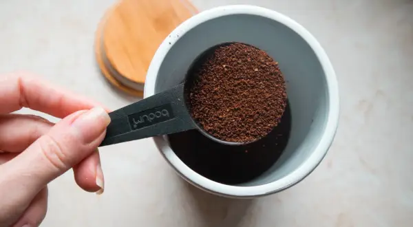 Eine Hand hält einen Löffel mit frisch gemahlenem Kaffeepulver, bereit, es in einen Behälter zu geben. Die Frage ist: wie viel Kaffeepulver für den perfekten Morgen?