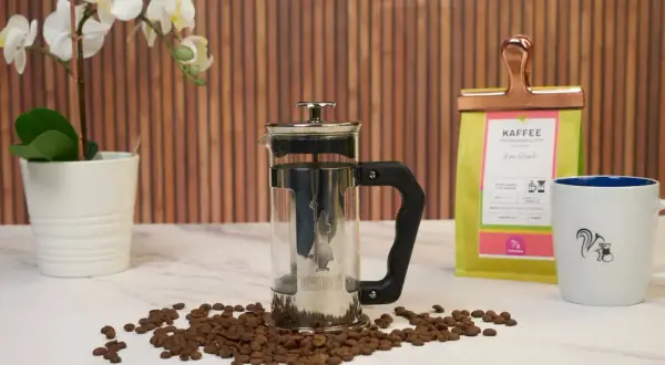 French press mit Kaffeesatz, bereit zum Entsorgen. Daneben Kaffeebohnen, eine Kaffeetüte und eine Tasse.