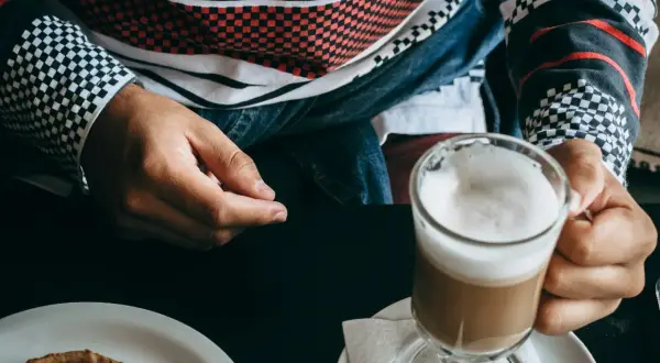 Person hält ein Glas mit schaumigem, kaltem Cappuccino. Auf dem Tisch liegt ein kleines Gebäck.