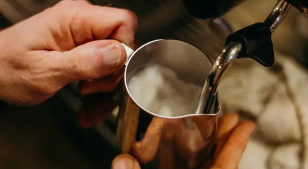 Hände halten Kännchen, während Dampf für perfekten Milchschaum wie aus der French Press aufsteigt.